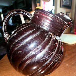 May include: A dark red glass pitcher with a swirl design. The pitcher has a handle and a spout.