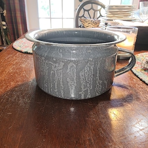 Op de afbeelding: Een grijze emaillekan met een brede, ronde basis en een enkele handgreep. De kan heeft een gespikkeld, getextureerd oppervlak.