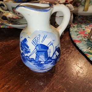 May include: A white ceramic pitcher with a blue and white windmill design. The pitcher has a handle and a spout.