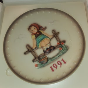 May include: A decorative plate featuring a young girl leaning on a wooden fence, with a basket and flowers. The girl wears a red headscarf, yellow top, and blue skirt. The plate is bordered with stars and the year 1991.