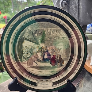 May include: A round decorative plate with a dark green border and gold trim. The centre features a detailed illustration of a festive indoor gathering with multiple figures. A small rectangular label at the bottom reads Christmas Eve at Mr. Wardle's.