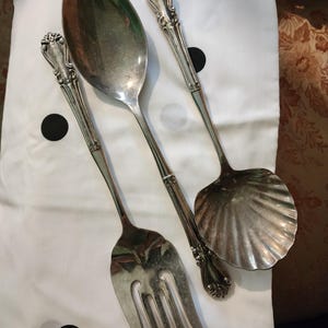 Op de afbeelding: Zilveren bestekset met een grote lepel, opscheplepel en vork. De gebruiksvoorwerpen hebben sierlijke handvatten en zijn gerangschikt op een witte doek met zwarte stippen. De opscheplepel heeft een schelpvormige kom.
