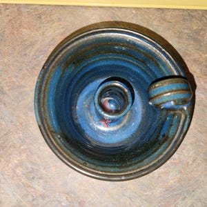 May include: A handmade ceramic candle holder with a deep blue glaze. The holder features a circular base with a central well for the candle and a small handle. The blue glaze has darker streaks, giving it a unique, artistic look.