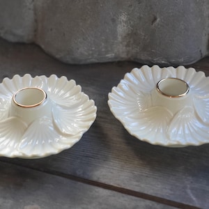 May include: Two off-white candle holders with a scalloped, shell-like design. Each holder has a central well with a gold-colored rim. The holders are placed on a wooden surface, suggesting a decorative or home decor theme.