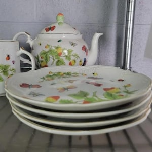 Könnte beinhalten: Ein Teeservice aus weißem Porzellan bestehend aus einer Teekanne, einer Tasse und einem Stapel von vier Tellern. Jedes Teil ist mit einem bunten Muster aus Erdbeeren, grünen Blättern, verschiedenen Schmetterlingen und kleinen Marienkäfern verziert.