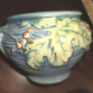 May include: A small, blue ceramic bowl with a leaf and grape design. The bowl features a textured surface and a light green and yellow leaf with brown grapes. The bowl has a small foot and a handle on one side.