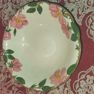 Puede incluir: Un cuenco de cerámica blanca con borde festoneado, adornado con diseños florales rosas y hojas verdes. El borde tiene un ribete dorado. El cuenco se utiliza probablemente para servir alimentos.