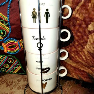 Puede incluir: Un juego de cuatro tazas apilables blancas con gráficos y texto en negro y dorado. Las tazas se sostienen en un soporte de metal cromado. El texto incluye "Female", "Girl Power" y otras palabras inspiradoras.