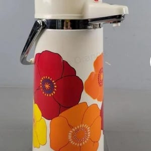 May include: Vintage cream-colored thermos with a floral design in red, orange, and yellow. The thermos has a white top with a red button and a silver handle. The floral pattern is a retro design.