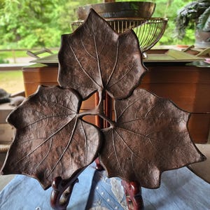 Pode incluir: Um prato decorativo em forma de folha, na cor bronze. O prato é suportado por um suporte de madeira escura. O design da folha apresenta veios detalhados. O prato é exibido sobre um tecido azul claro com bordados de folhas brancas.