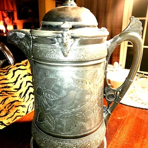 Puede incluir: Una jarra de plata con un diseño ornamentado y una tapa. La jarra tiene un patrón floral y un asa con un remate decorativo. La jarra está sentada sobre una mesa de madera.