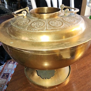 Puede incluir: Una olla caliente grande, redonda y de latón con tapa y asas decorativas. La olla tiene una base elevada y diseños intrincados en la tapa.