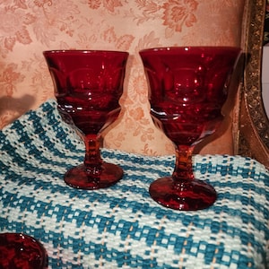 Puede incluir: Dos copas de vino de color rojo rubí con un diseño texturizado. Las copas se exhiben sobre una tela estampada en azul y blanco. El fondo presenta una tela con estampado floral.