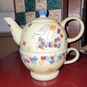 Könnte beinhalten: Gelbes Keramik-Teeservice, bestehend aus einer Teekanne und einer Tasse. Die Teekanne hat einen blauen Griff und Deckel, die Tasse ist darunter gestapelt. Beide Teile sind mit einem Blumen- und Engeldesign in mehreren Farben verziert.