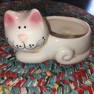 May include: White ceramic cat planter with pink ears and black eyes. The planter has a round opening on the back for planting.