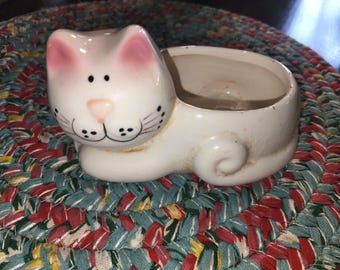 May include: White ceramic cat planter with pink ears and black eyes. The planter has a round opening on the back for planting.