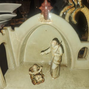 Puede incluir: Un belén de cerámica en color beige y marrón. La escena representa a un bebé Jesús en un pesebre, un pastor con un bastón y una palmera. Las palabras "Paz" están inscritas en la parte superior de la estructura.