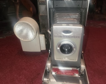 Puede incluir: Una cámara Polaroid Land vintage con cuerpo negro y ribete plateado. La cámara tiene un flash incorporado y una lente con un anillo plateado. La cámara está abierta y el paquete de película es visible. La cámara tiene el texto "POLAROID ELECTRIC EYE" impreso en la parte delantera.