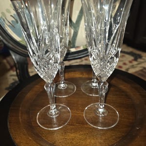 Könnte beinhalten: Ein Set aus vier Sektgläsern aus klarem Glas mit komplexen vertikalen Schliffmustern und einem goldfarbenen Rand. Jedes Glas hat einen schlanken Stiel und einen runden Fuß und zeigt ein facettiertes Design, das das Licht reflektiert.