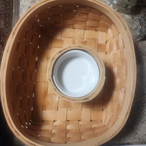 May include: Oval woven basket with a central, circular white ceramic bowl. The basket is made of light brown woven strips, creating a textured surface. The bowl is centered within the basket, offering a contrasting white interior.