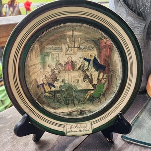 May include: A round decorative ceramic plate featuring a colourful illustration of a group of men gathered around a table, with one man standing and gesturing. The plate has a dark green rim and a label at the bottom reading Mr. Pickwick addresses the Club.