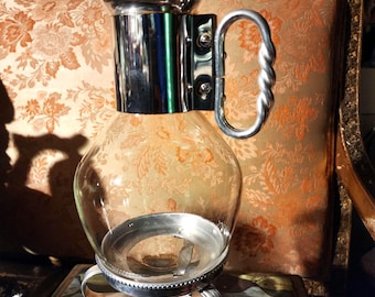 Pode incluir: Uma cafeteira de vidro transparente e metal prateado, com um corpo de vidro redondo e uma alça prateada. A cafeteira tem uma base prateada e uma tampa com uma pequena alça. O fundo inclui tecido estampado e obras de arte emolduradas.