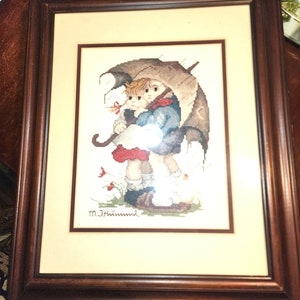 May include: Framed cross-stitch artwork featuring two children under an umbrella in a rainy setting. The artwork is signed "M.J.Hummel" in the bottom right corner.