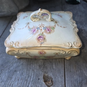 May include: A vintage porcelain trinket box with a lid. The box is cream-colored with gold trim and feet. The lid features a floral design in pink, purple, and blue. The handle is gold-colored.