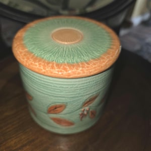 May include: A cylindrical ceramic jar with a lid, featuring a light green crackle glaze finish. The lid has a peach-colored rim and a central circular detail. The body of the jar is decorated with embossed orange leaf and berry motifs.