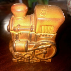 Puede incluir: Una jarra de galletas de cerámica marrón con forma de locomotora. La jarra tiene la palabra "Cookies" escrita en la parte superior.