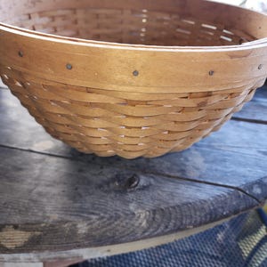 May include: A round, woven basket crafted from light brown wood. The basket features a wide, open top and a gently curved, textured body. Small metal fasteners secure the rim. The basket rests on a weathered wooden surface.
