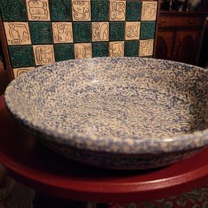 Puede incluir: Un cuenco grande y redondo de cerámica con un patrón moteado azul y blanco. El cuenco está sobre una superficie roja, con un panel decorativo de azulejos de estilo maya en el fondo. El diámetro del cuenco es de unos 30 cm.