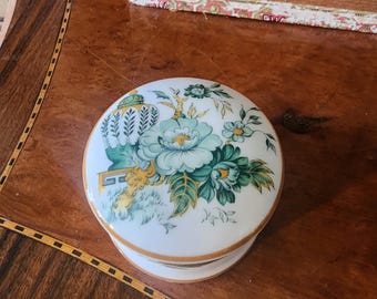 Könnte beinhalten: Eine weiße Porzellan-Schmuckdose mit Blumenmuster. Der Deckel zeigt ein grünes und gelbes Blumenmuster auf weißem Hintergrund. Die Dose ist rund und hat einen goldenen Rand.
