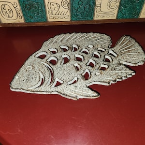 May include: A decorative, fish-shaped trivet with a distressed, textured finish. The trivet is primarily gray with white accents and cut-out details. It rests on a red surface, with a glimpse of a decorative wooden panel in the background.