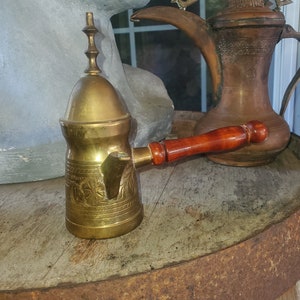 Puede incluir: Una cafetera de latón con un diseño ornamentado y un mango de madera. La cafetera tiene una tapa abovedada con un remate en la parte superior. La cafetera está sobre una superficie de madera.