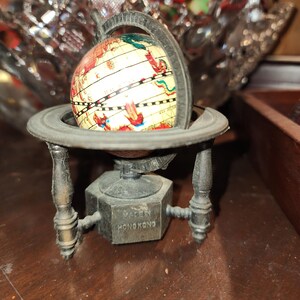Puede incluir: Un pequeño globo terráqueo sobre un soporte decorativo de metal. El globo presenta un mapa con detalles rojos, amarillos y verdes. El soporte es de color gris oscuro con una base hexagonal y el texto "MADE IN HONGKONG".