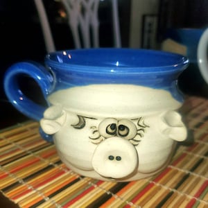 May include: A white ceramic mug with a blue rim and a whimsical pig face design. The mug has a handle and features black eyes, a snout, and two button-like nostrils.