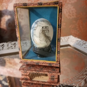 Puede incluir: Objeto decorativo en forma de huevo con una escena de tigre pintada, exhibido en una vitrina de cristal con fondo azul. La vitrina tiene un marco decorativo y se apoya sobre una superficie espejada. El huevo está montado sobre un soporte negro.