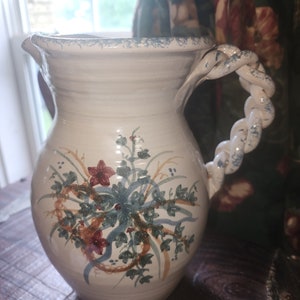 Puede incluir: Una jarra de cerámica blanca con asa trenzada y un diseño floral en tonos rojos, verdes y azules.