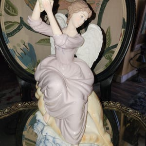 May include: A porcelain figurine of an angel with light brown hair and white wings, wearing a pale lavender dress. The angel is seated on a large, tan seashell atop stylized blue and white waves, holding a smaller conch shell above their head.