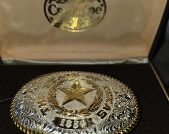 May include: Silver and gold-tone Texas state belt buckle with a star in the center. The buckle has an oval shape with ornate scrollwork and the words "THE STATE OF TEXAS" and "1836" engraved on it. The buckle is in a presentation box.
