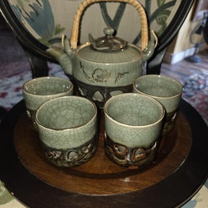 Puede incluir: Un juego de té de cerámica compuesto por una tetera con asa trenzada y cuatro tazas a juego. Las piezas presentan un acabado de esmalte craquelado verde claro en la parte superior y una base decorativa calada en marrón oscuro con motivos florales.