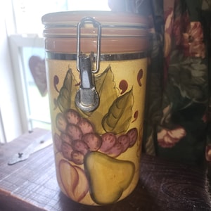 Könnte beinhalten: Eine Keramikdose mit braunem Deckel und einem Metallverschluss. Die Dose ist mit einem handgemalten Design aus Trauben, Birnen und Blättern in Grün-, Gelb- und Lilatönen verziert.