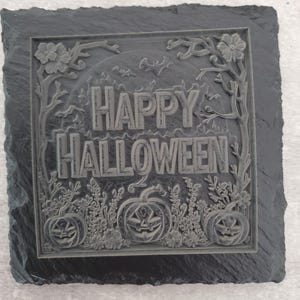 May include: A dark gray slate sign with the words "HAPPY HALLOWEEN" carved into the surface. The design includes carved pumpkins, flowers, and bats, creating a festive Halloween theme.