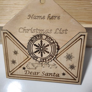 May include: Wooden Christmas list holder shaped like an envelope. The holder has the words "Name here", "Christmas List", and "Dear Santa" engraved on it. A circular emblem with "North Pole Post Office" is in the center.