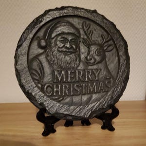 Op de afbeelding: Een donkergrijze, ronde kerstversiering van leisteen met een reliëf van de kerstman en een rendier, met de tekst "MERRY CHRISTMAS". De leisteen is gemonteerd op een zwarte standaard, geschikt voor weergave op een plank of tafel.