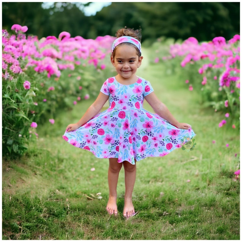 May include: A young girl wearing a light blue dress with a floral print stands in a field of pink flowers. She is wearing a headband with a floral design.