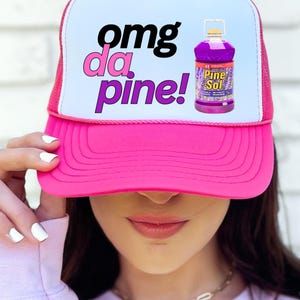 May include: A pink trucker hat with a white front panel featuring the text "omg da pine!" in black and pink, and an image of a Pine-Sol bottle. The hat has a pink brim and a pink mesh back.