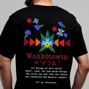 May include: Black t-shirt with a colourful graphic design featuring flowers, geometric shapes, and the text "Wahkôtowin <"d"j"d>." The text below the graphic reads "All beings of this world: water, land, sky and earth beings~ who exist now and into the future are connected and deserve respect. All my relations."