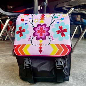May include: A pink backpack with a floral pattern and black straps. The floral pattern features pink, red, yellow, and blue flowers. The backpack has a black zipper and a black bottom.